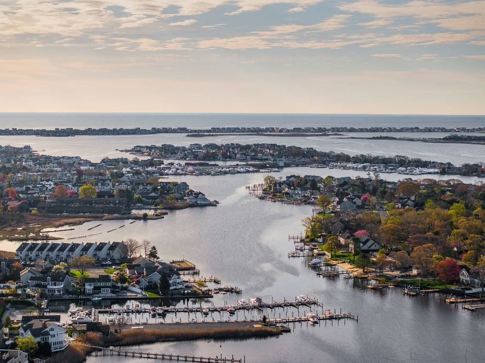 Aerial view of a coastal inlet with houses, docks, and boats surrounded by water and trees under a cloudy sky. Ocean County New Jersey from an aerial view