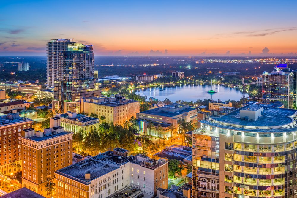 Aerial view of Orlando, Florida at dusk with a lake in the center, illuminated buildings, and a colorful sky.