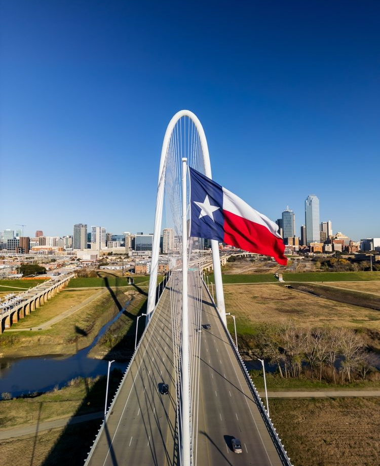 Texas flag on a bridge over a river with a city skyline under a clear blue sky