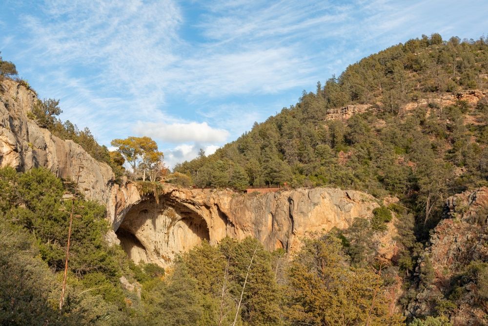 Rocky canyon with a natural stone arch under a blue sky, surrounded by trees and hills.Tonto Natural Bridge State Park. Autumn.