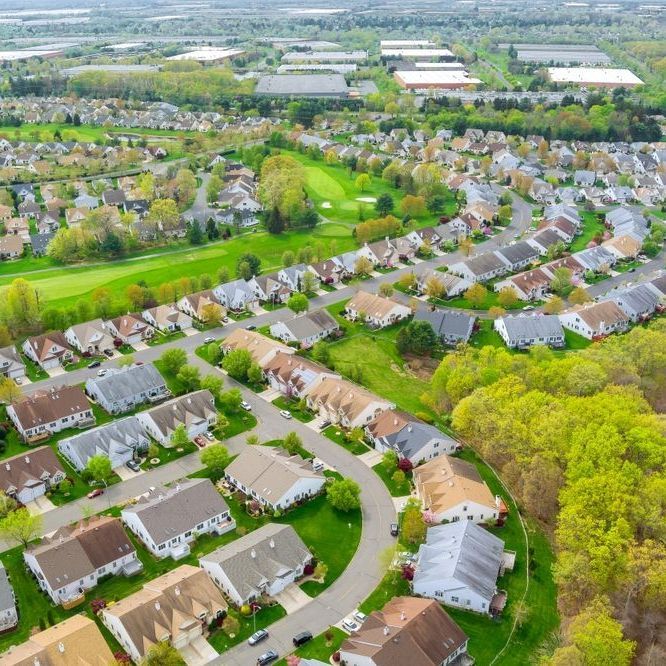 Aerial view of a suburban neighborhood with houses, green lawns, and a golf course. Trees and blue sky visible in the background.