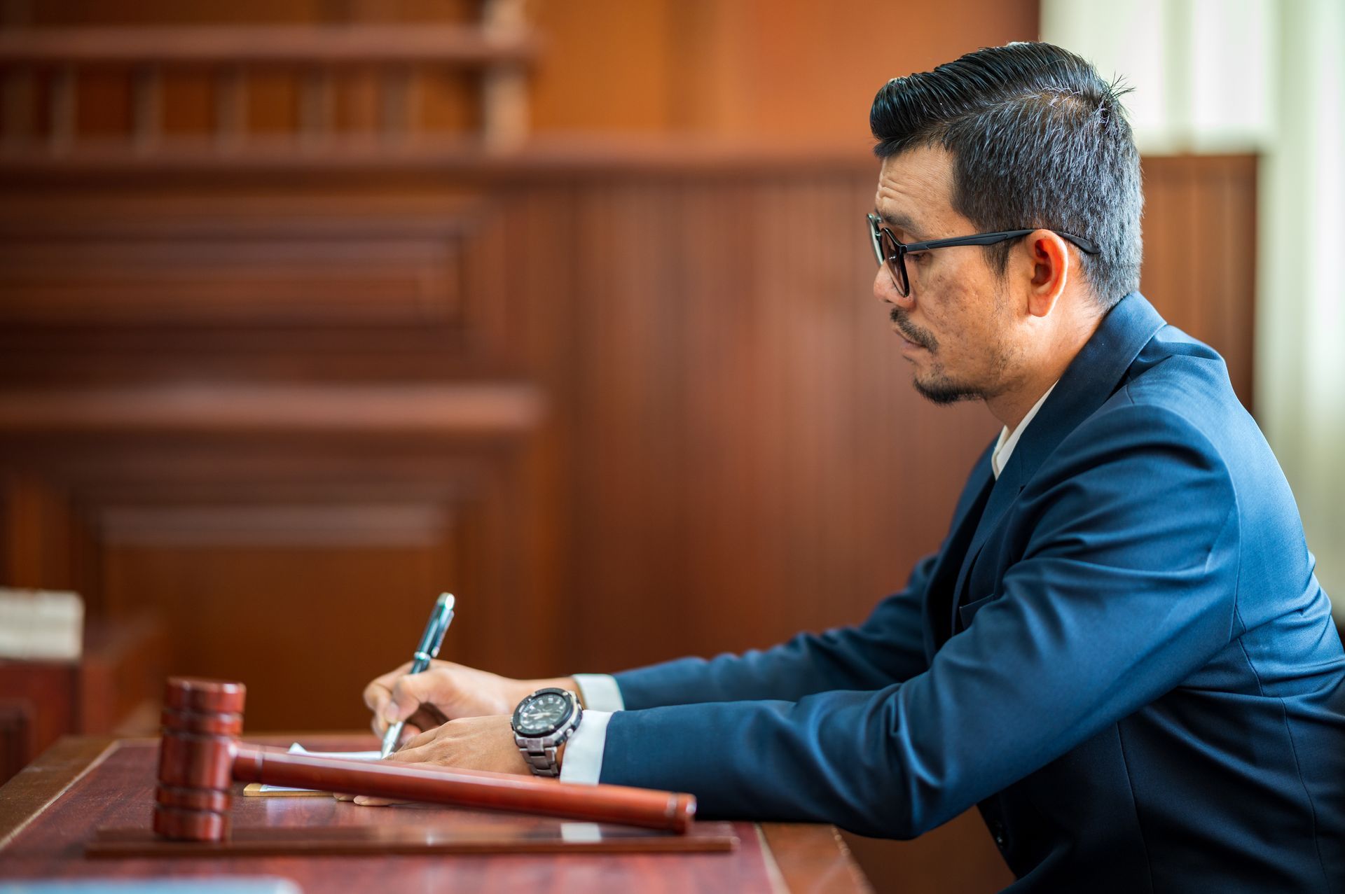 A person in a suit is seated at a wooden desk in a courtroom, writing with a pen. A gavel lies in front of them.