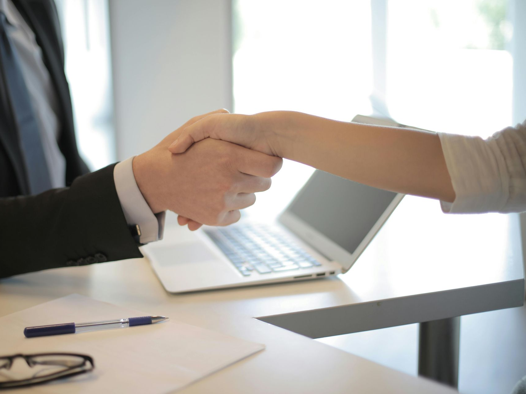 Two people shaking hands over a desk with a laptop, pen, glasses, and paper.