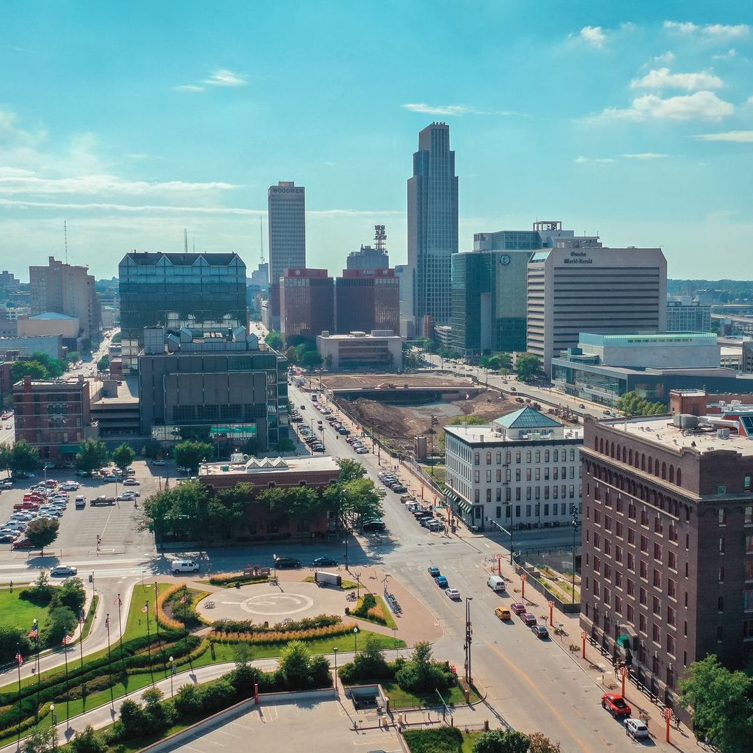Aerial view of downtown Omaha, Nebraska on a sunny day. Skyscrapers and buildings line the streets, with a park in the foreground.