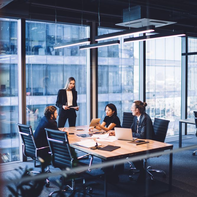 Business meeting in modern office, with four people seated around a table, and one standing.