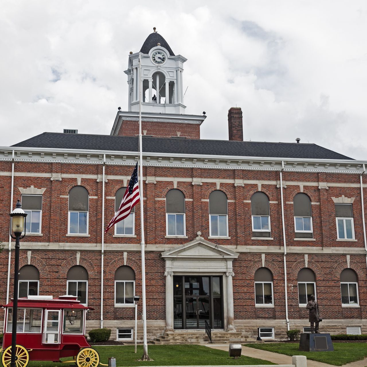 Brick courthouse with clock tower and American flag, in front of a small red and yellow wagon.