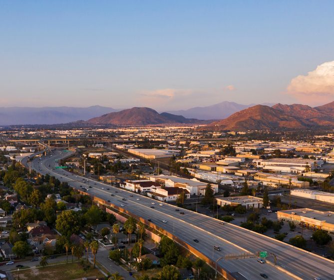 Aerial view of a highway winding through an industrial area and residential neighborhoods with mountains in the background under a blue sky.