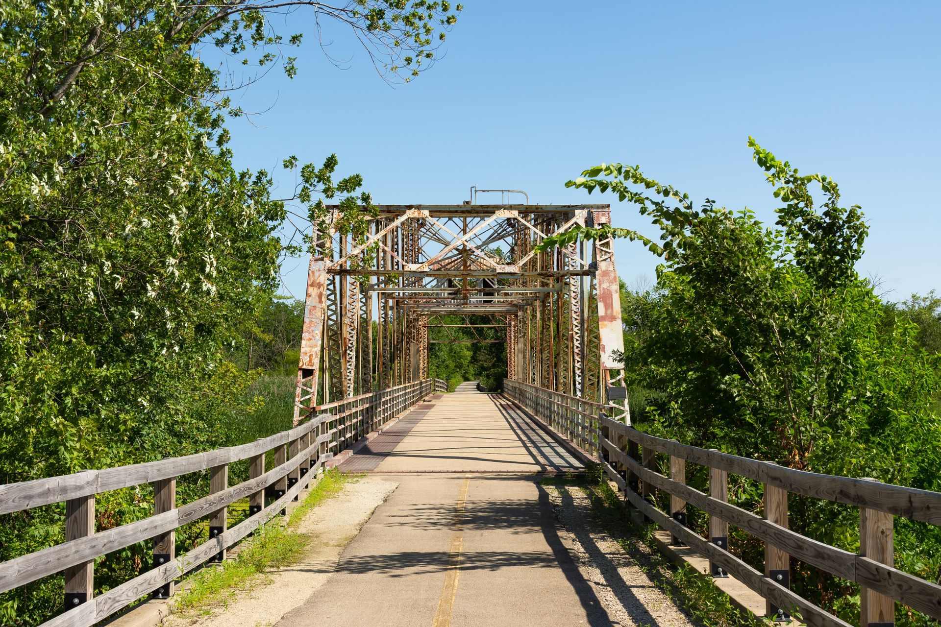 Bridge with a metal truss frame, over a paved path with wooden railings, surrounded by green trees under a clear blue sky.