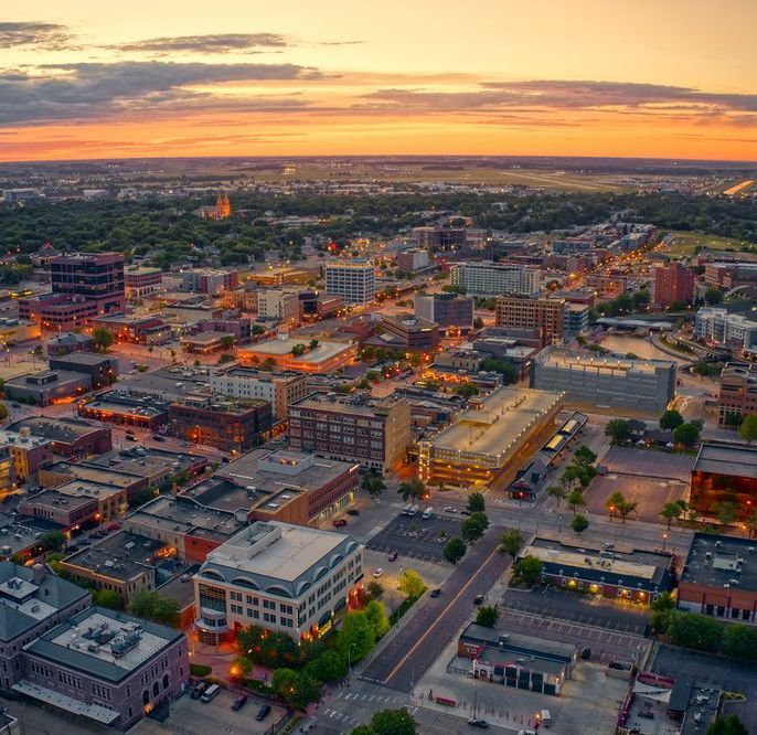 Aerial view of a city at sunset, buildings illuminated with warm light, a fiery orange sky in the background.