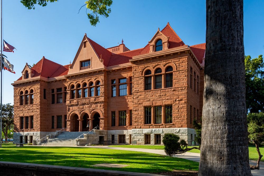 Red brick courthouse with a red roof on a sunny day. Flags fly in front of the building, and green grass surrounds it.