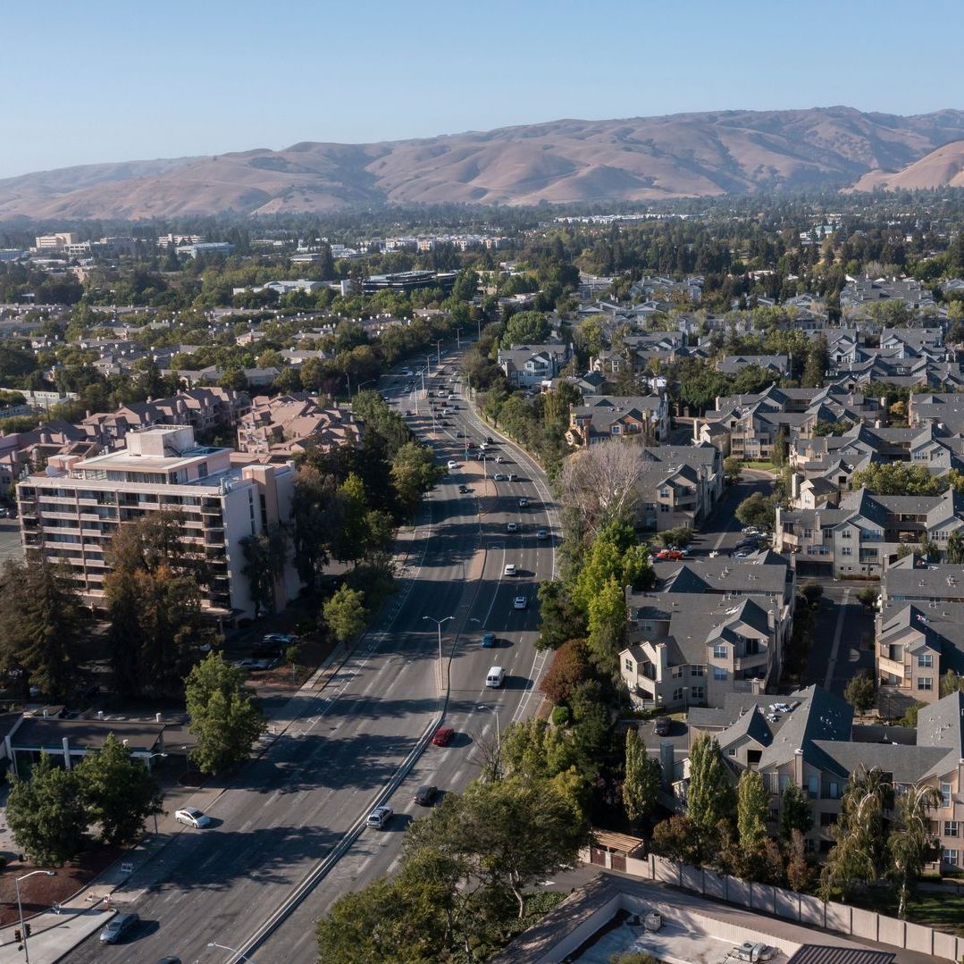 Aerial view of a suburban neighborhood with a road lined with trees, with mountains in the distance under a blue sky.
