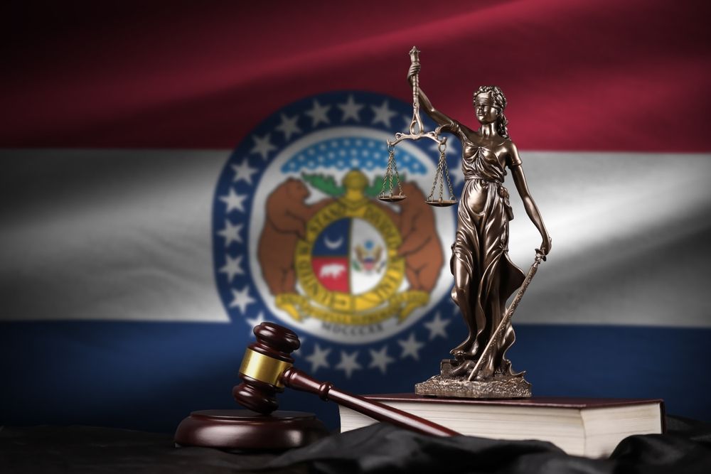 Statue of Lady Justice and gavel in front of Missouri state flag. The statue balances scales, representing law and justice.