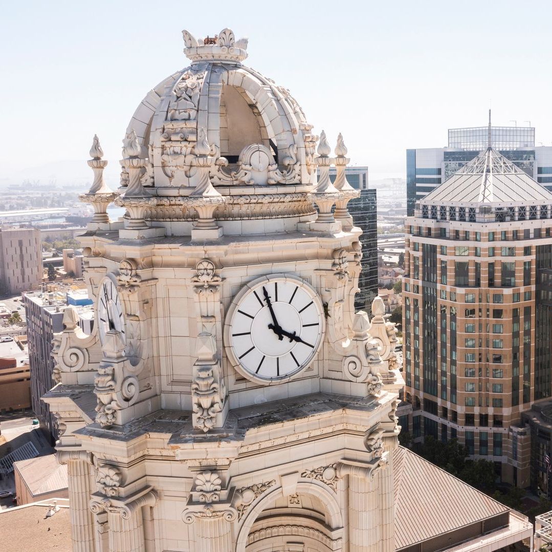 Clock tower with ornate white facade, surrounded by city buildings under a clear blue sky. The clock shows 2:47.