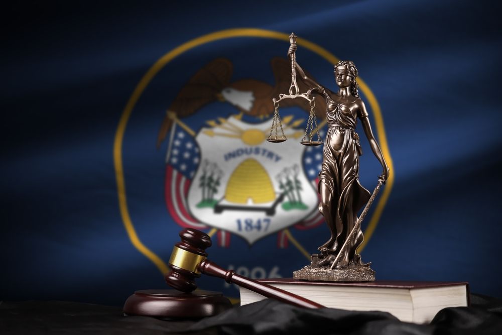 A statue of Lady Justice with scales and a gavel on a book against a backdrop of the Utah state flag.