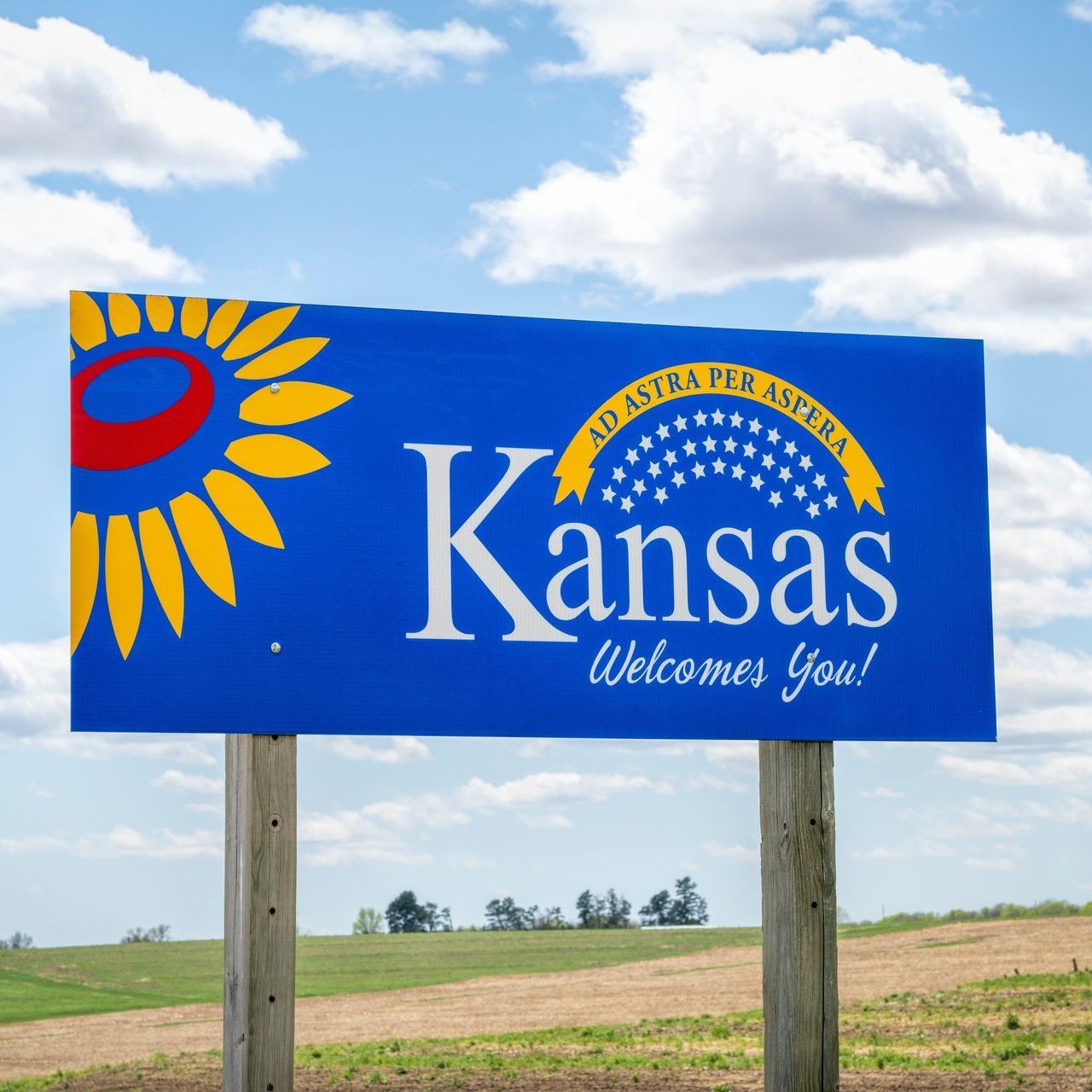 Kansas welcome sign with a blue background, sunflower, and state motto against a cloudy sky and field.