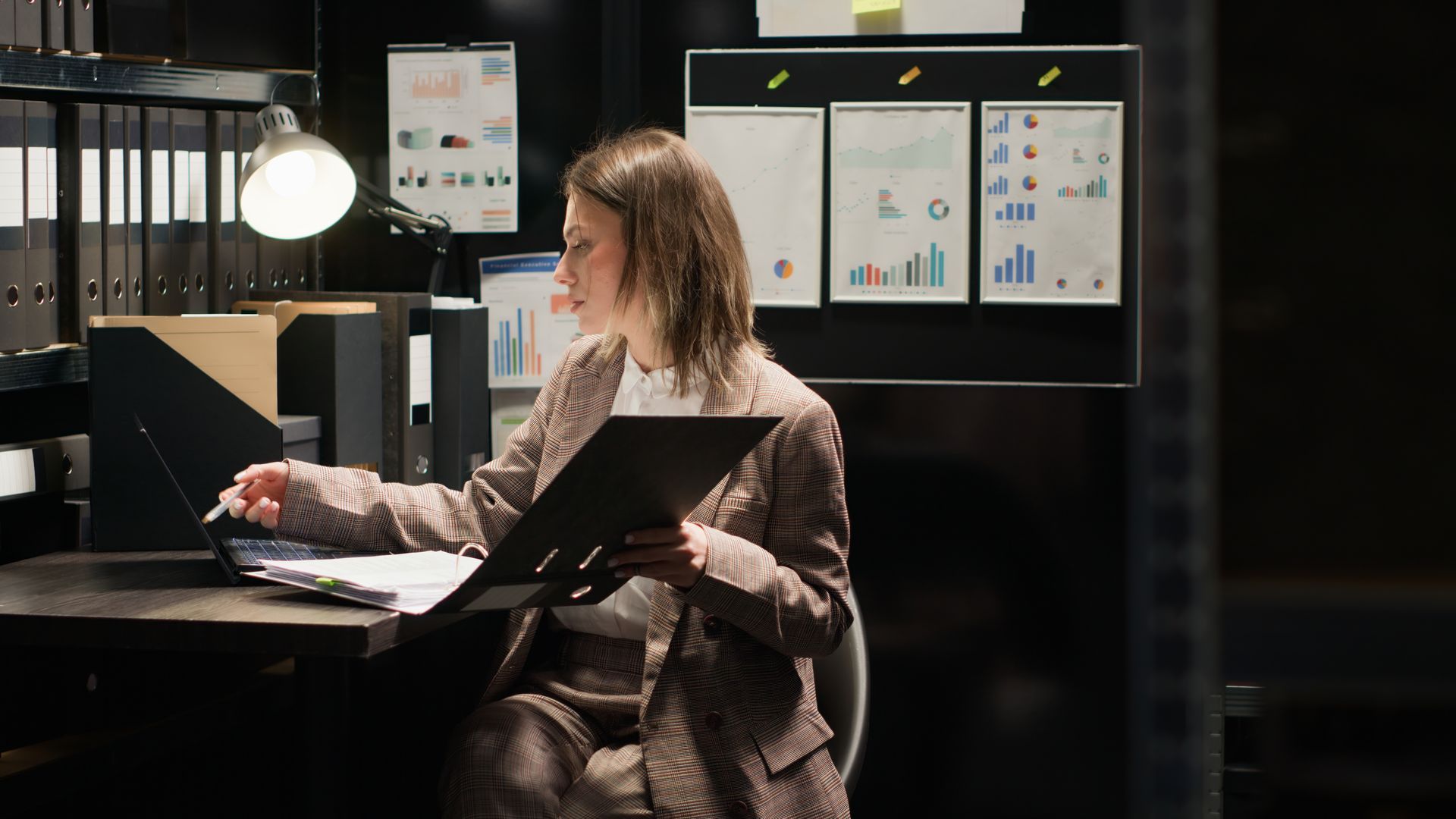 A woman is sitting at a desk in an office holding a clipboard and a laptop cross-referencing ongoing background checks