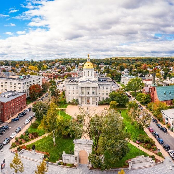 Aerial view of the New Hampshire State House in Concord, with a gold dome. Green lawns and trees surround the white building, under a cloudy sky.