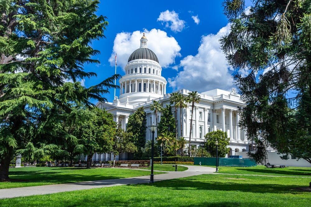 California State Capitol building, white with a dome, seen from a park with trees and a blue sky.