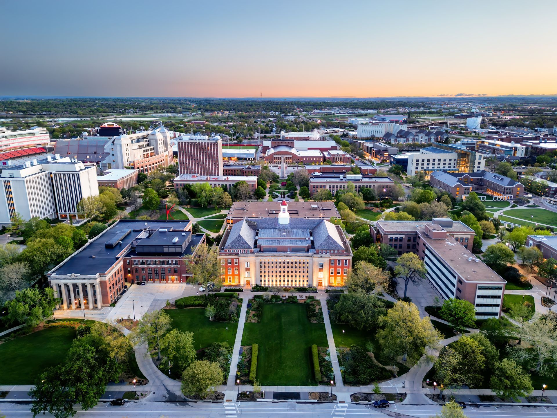 Aerial view of a university campus at dusk. Buildings are illuminated by warm light, surrounded by green lawns and trees.
