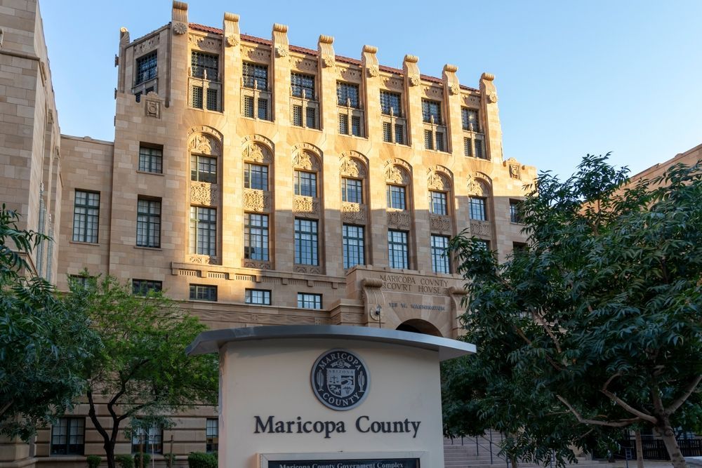 Maricopa County building exterior with sign in the foreground. Tan stone structure with arched windows, and a clear sky.