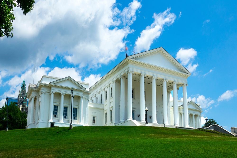 Virginia State Capitol building with white columns, on a green hill, against a blue sky with clouds.