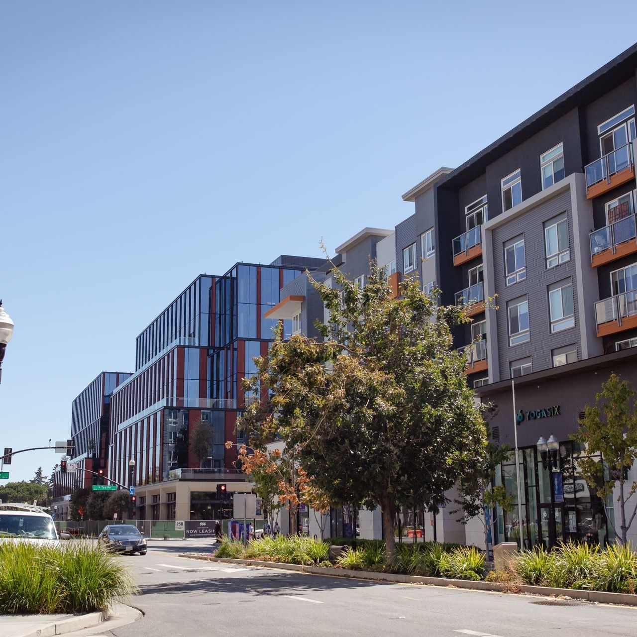 Street view of modern buildings with glass and dark facade, clear blue sky overhead. A car drives down the street, with greenery and trees.