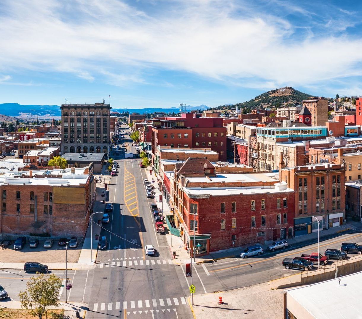 Aerial view of a downtown area with brick buildings, a street with cars, and a hill in the background under a blue sky.