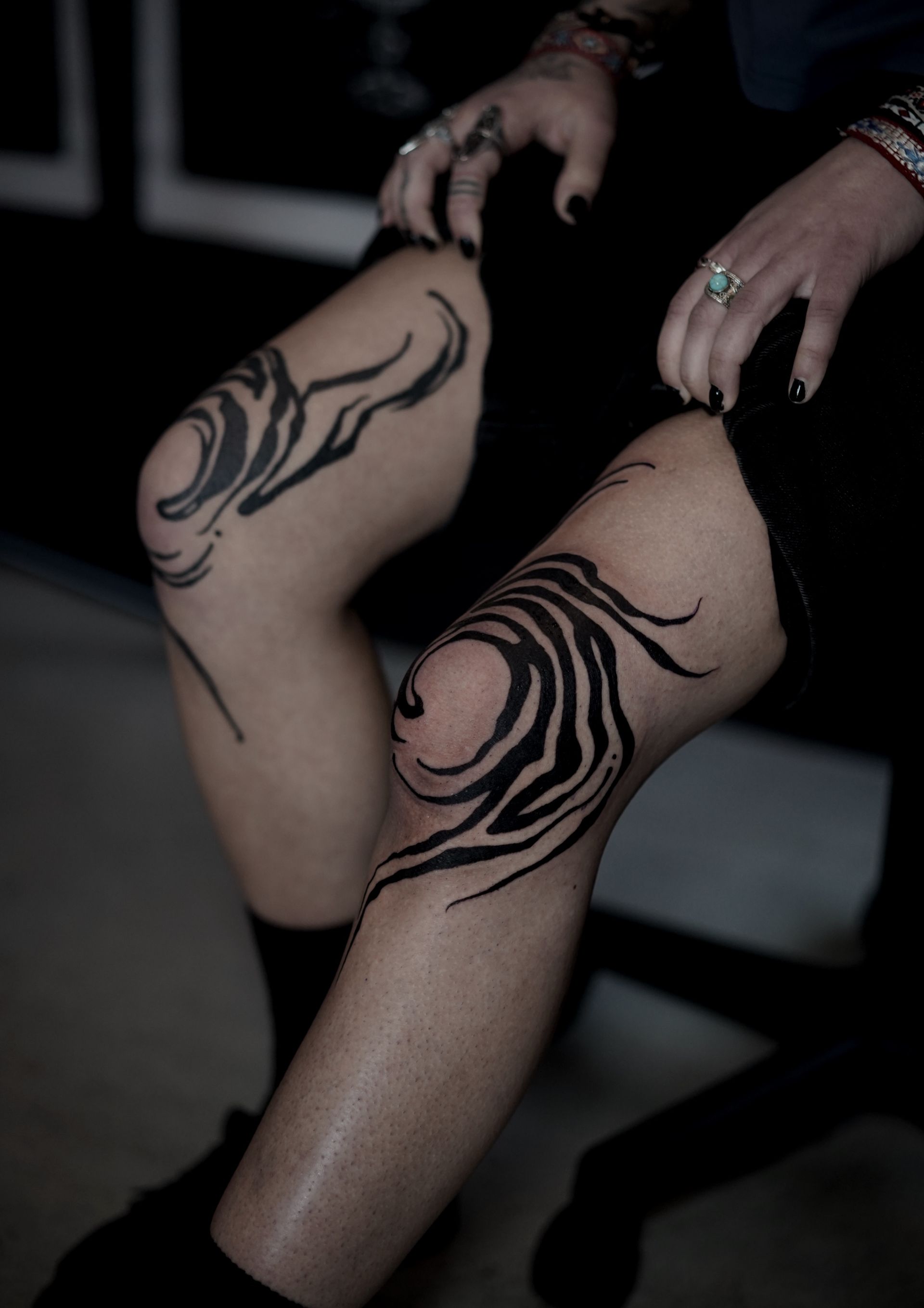 Person's knees with swirling black tribal tattoos. They wear black clothes and rings on their fingers.