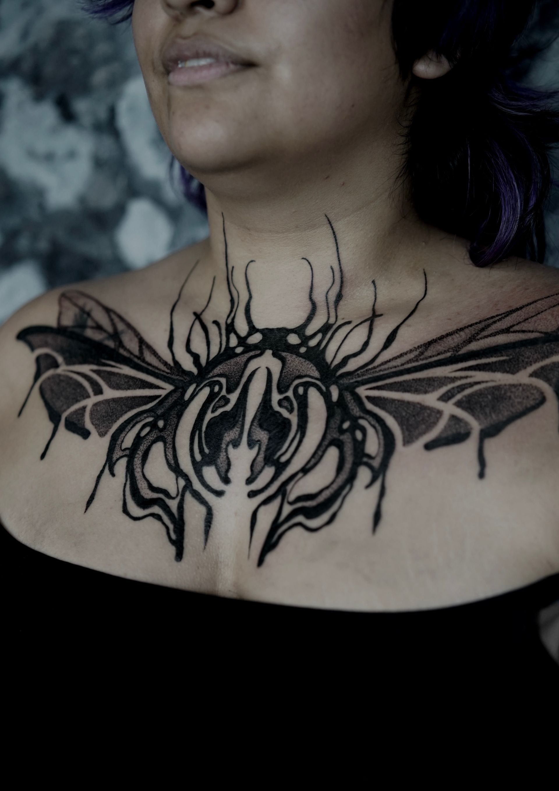 Woman with a black insect tattoo on her chest, against a dark background.