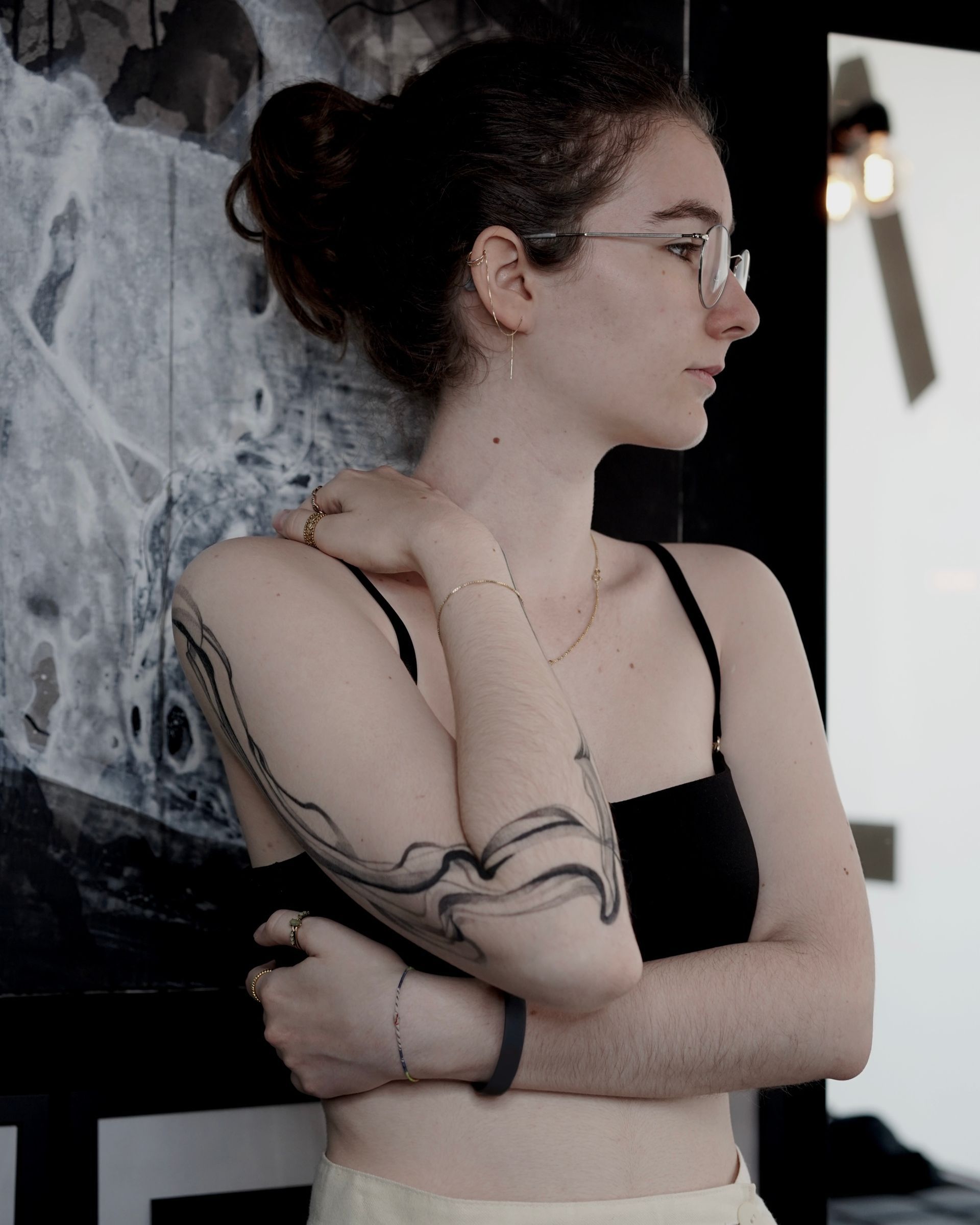 Woman with glasses and arm tattoo, leaning against a dark wall, looking to the right.