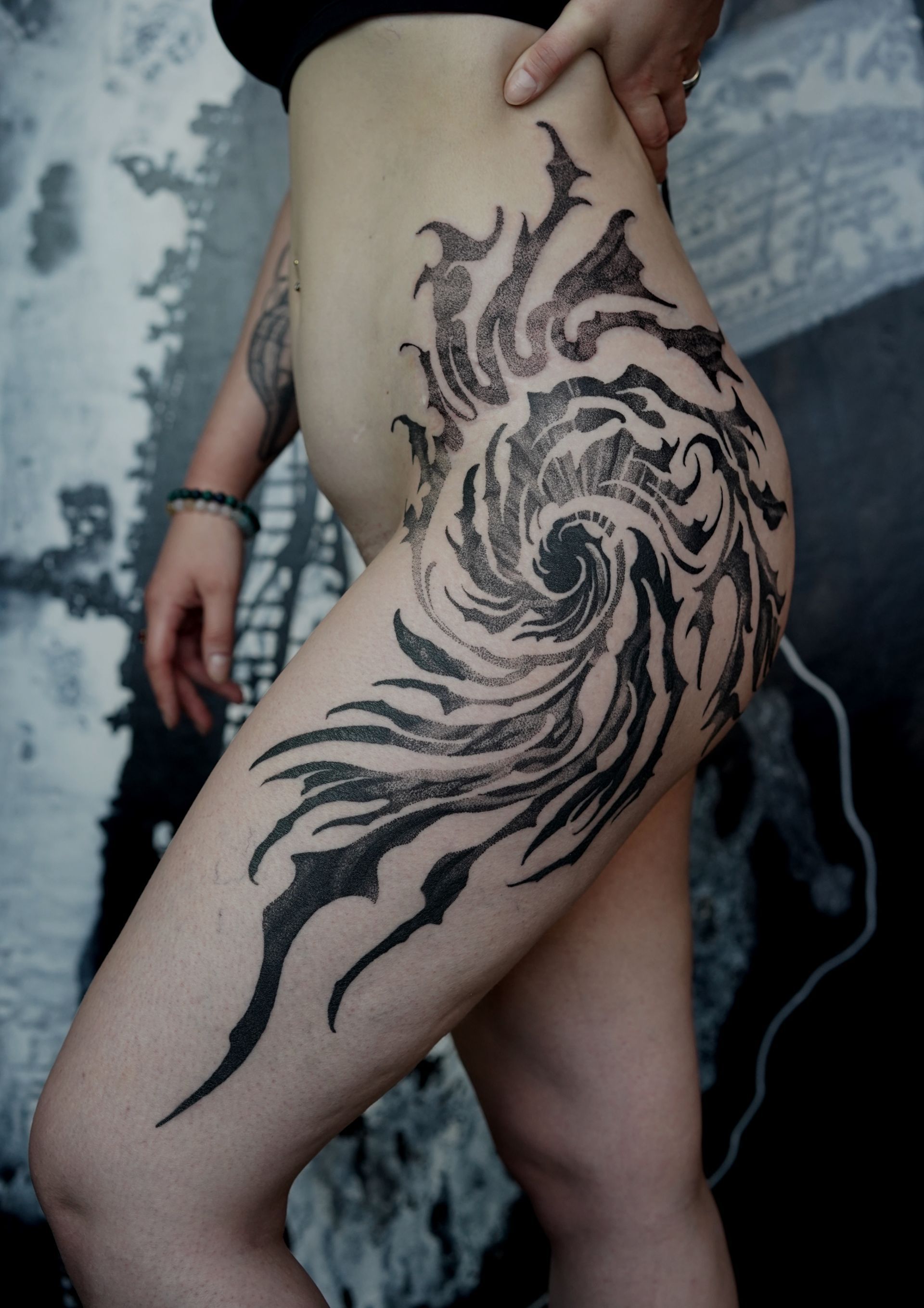 Black abstract swirling tattoo on a person's thigh and hip; set against a textured background.