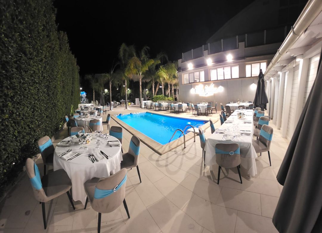 Zona pranzo serale a bordo piscina con tavoli bianchi, sedie blu e piscina illuminata.