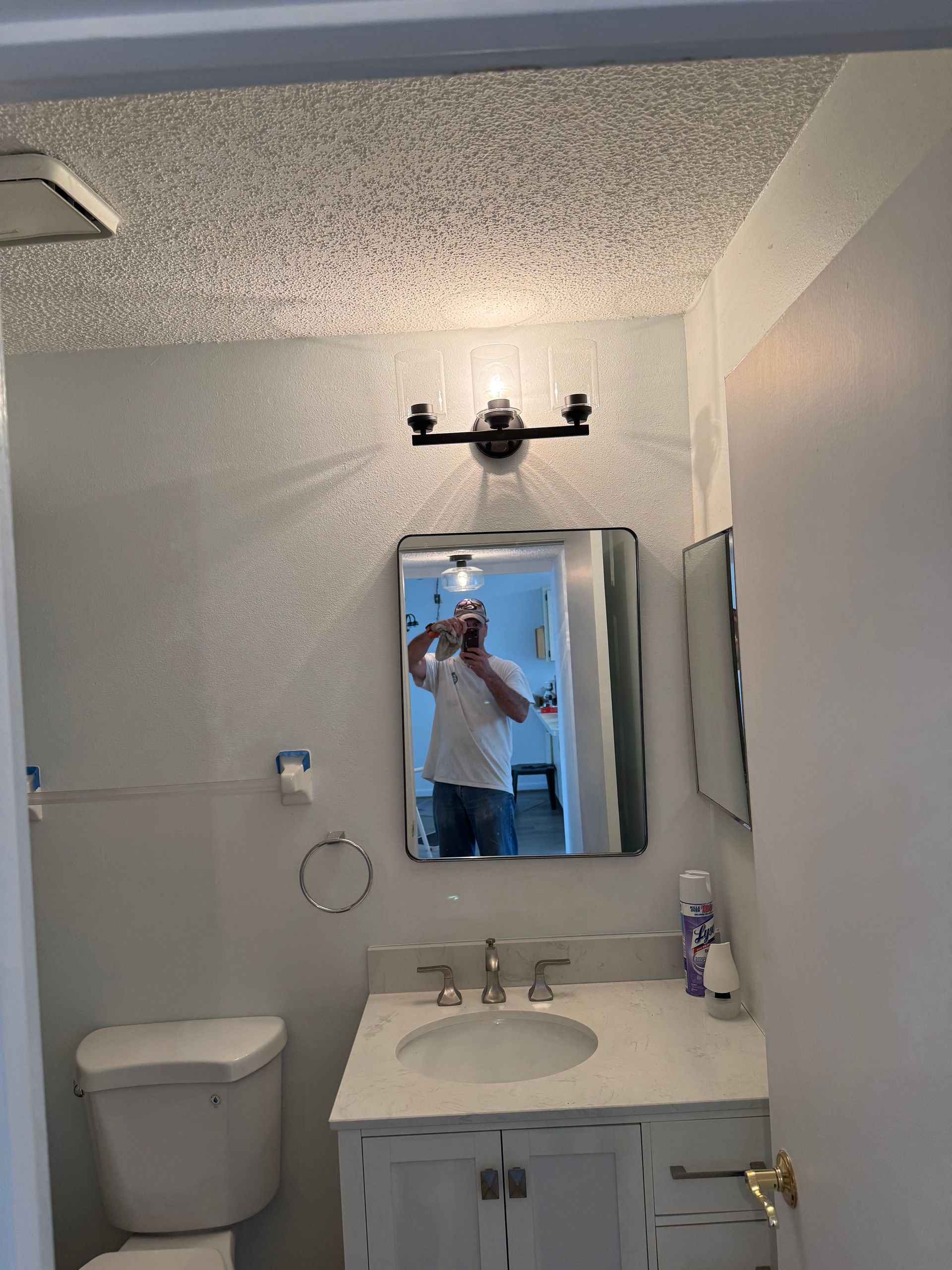 Small bathroom with toilet, sink, mirror, and wall light; a person is reflected taking the photo.