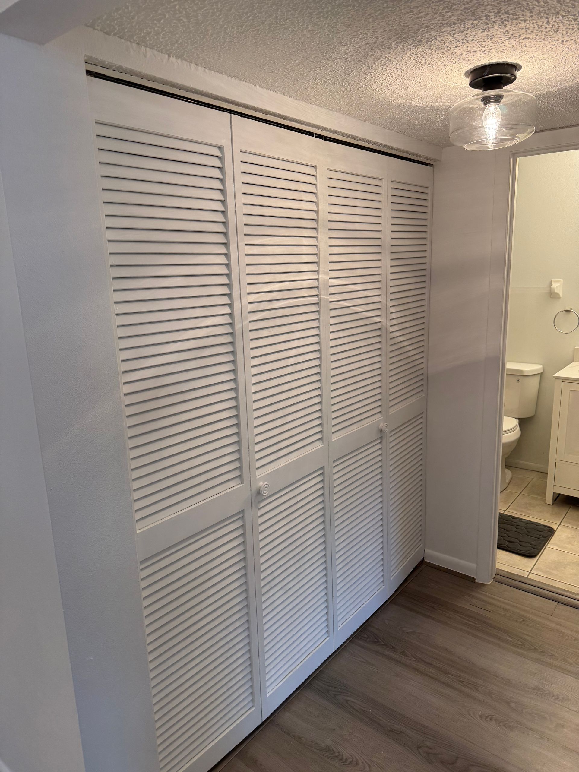 White louvered closet doors in a hallway beside a bathroom doorway with a light fixture.