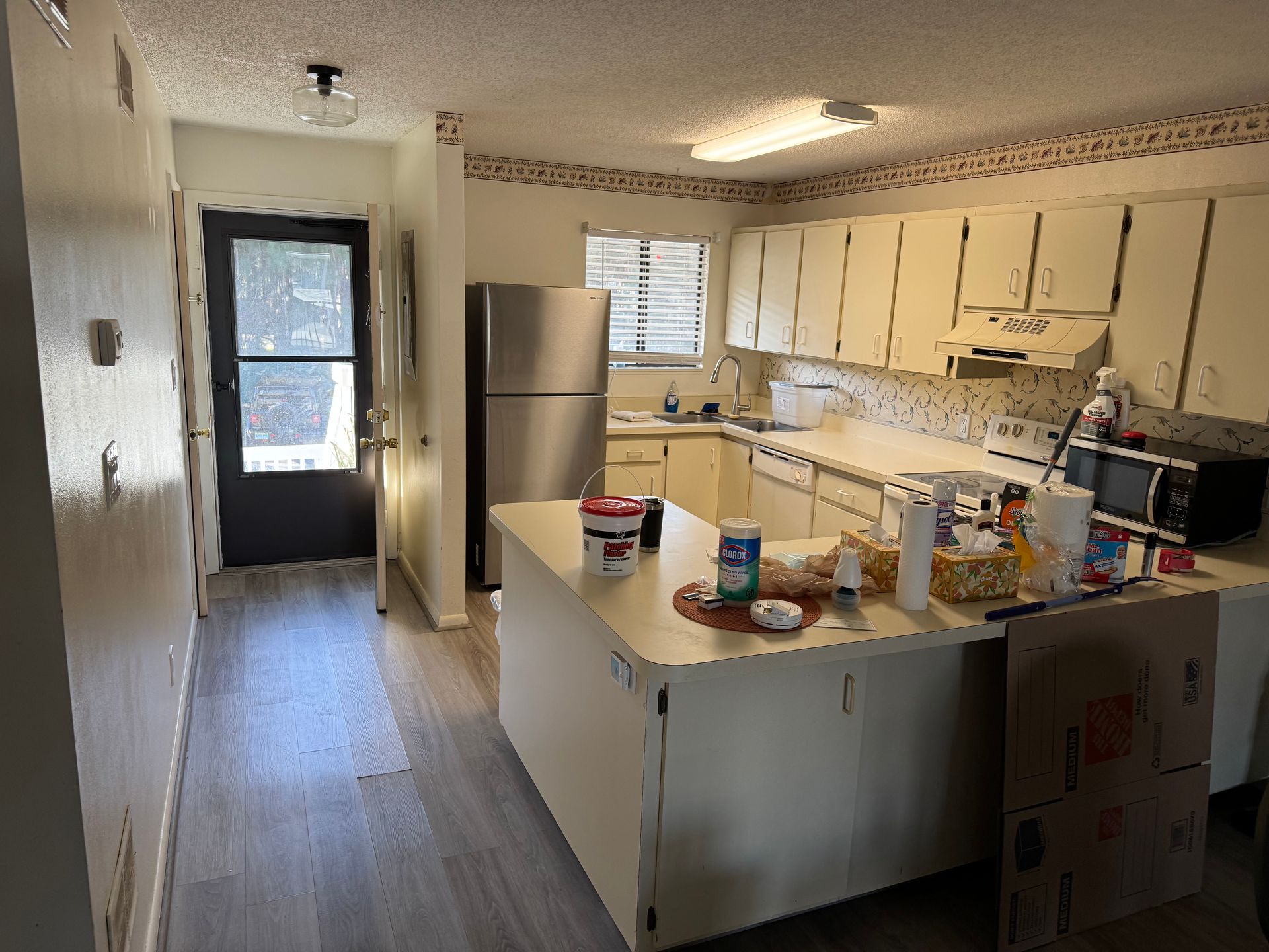 Small kitchen with white cabinets, stainless fridge, and a cluttered island beside a bright hallway.