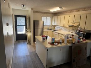 Kitchen with white cabinets, stainless fridge, island counter, and a dark exterior door in a narrow hallway.
