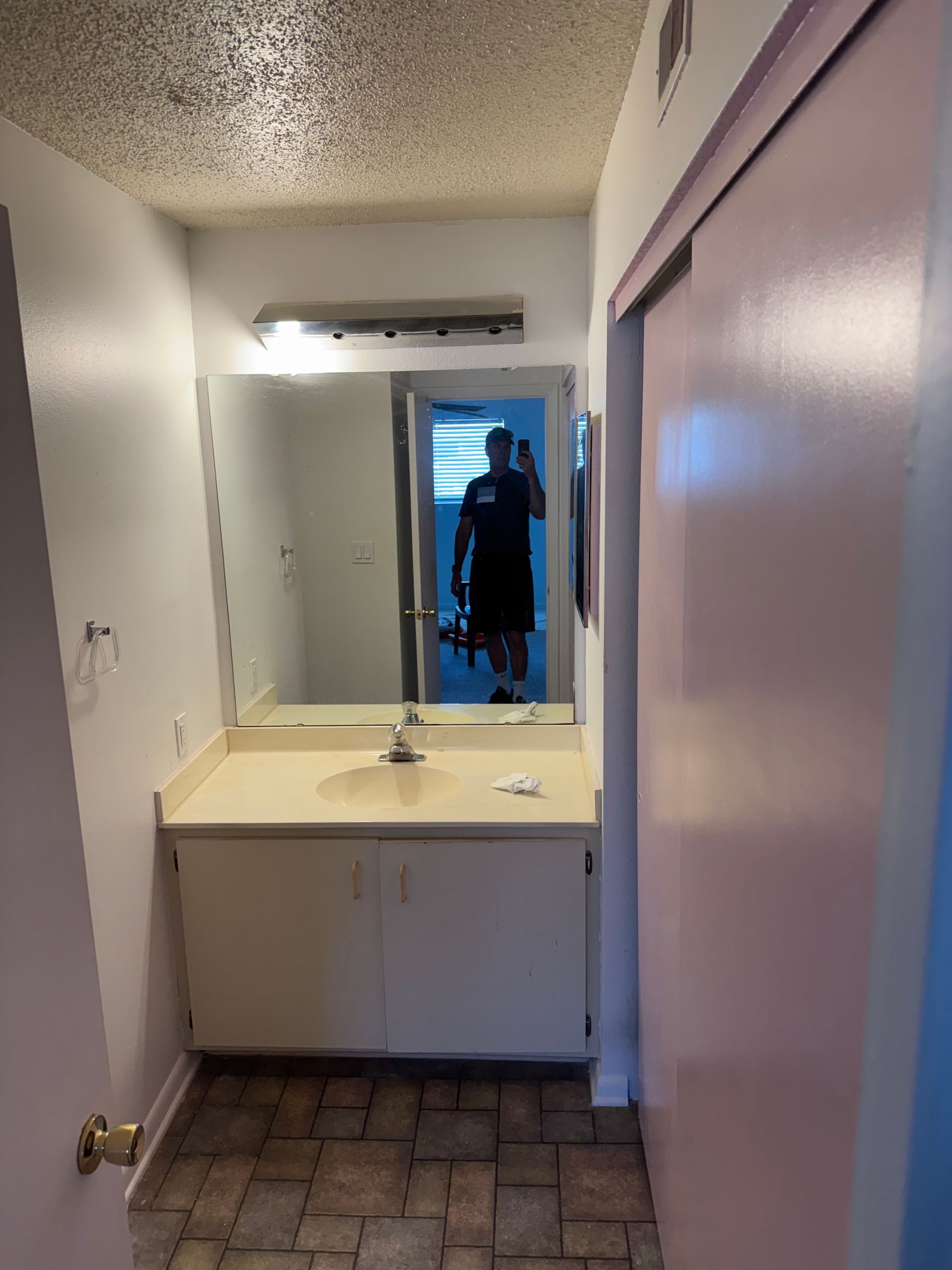 Narrow bathroom with white vanity, mirror, and a silhouetted person reflected in the doorway.