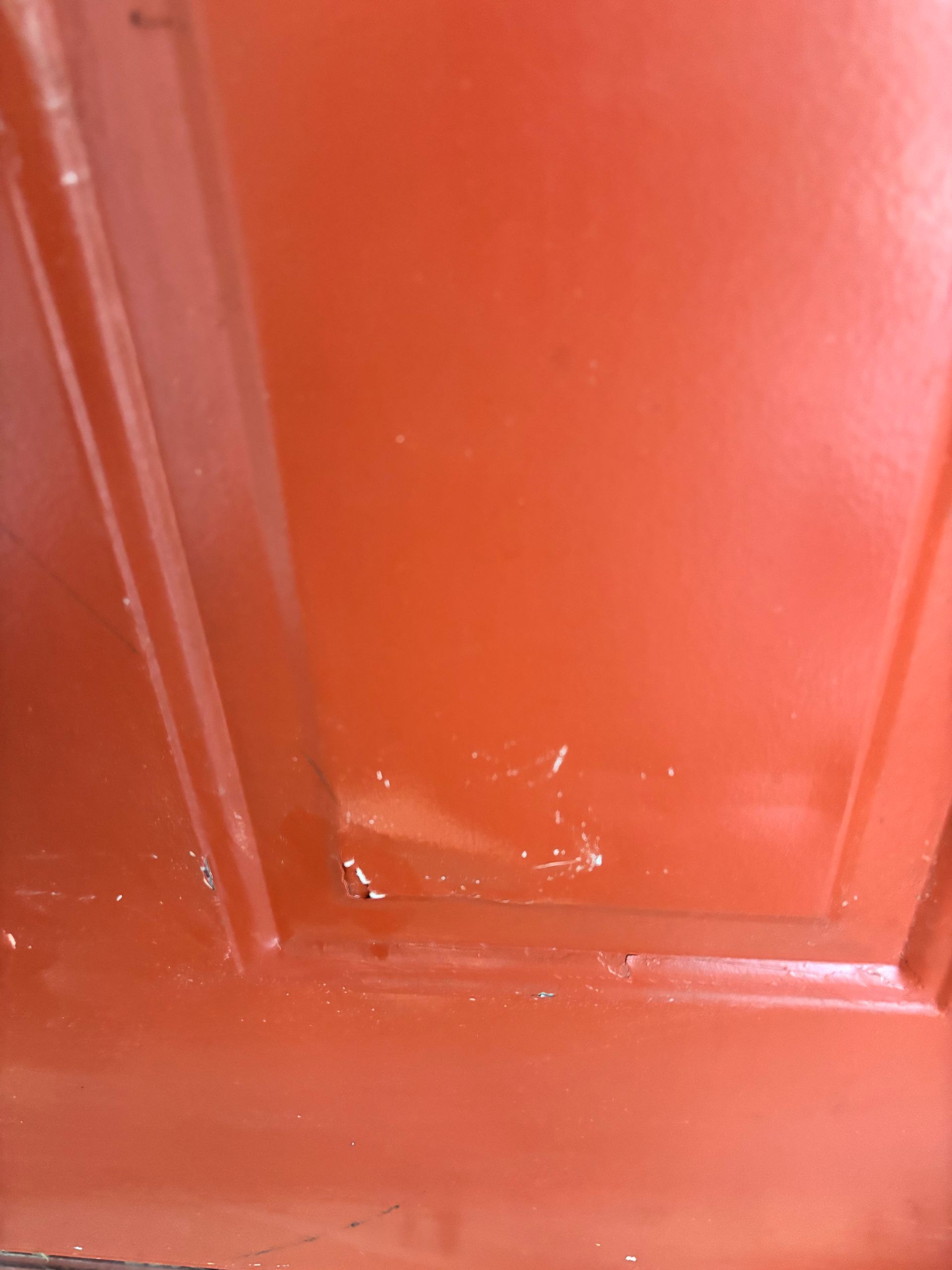 Close-up of a red painted metal door panel with recessed molding and a small dent near the bottom.