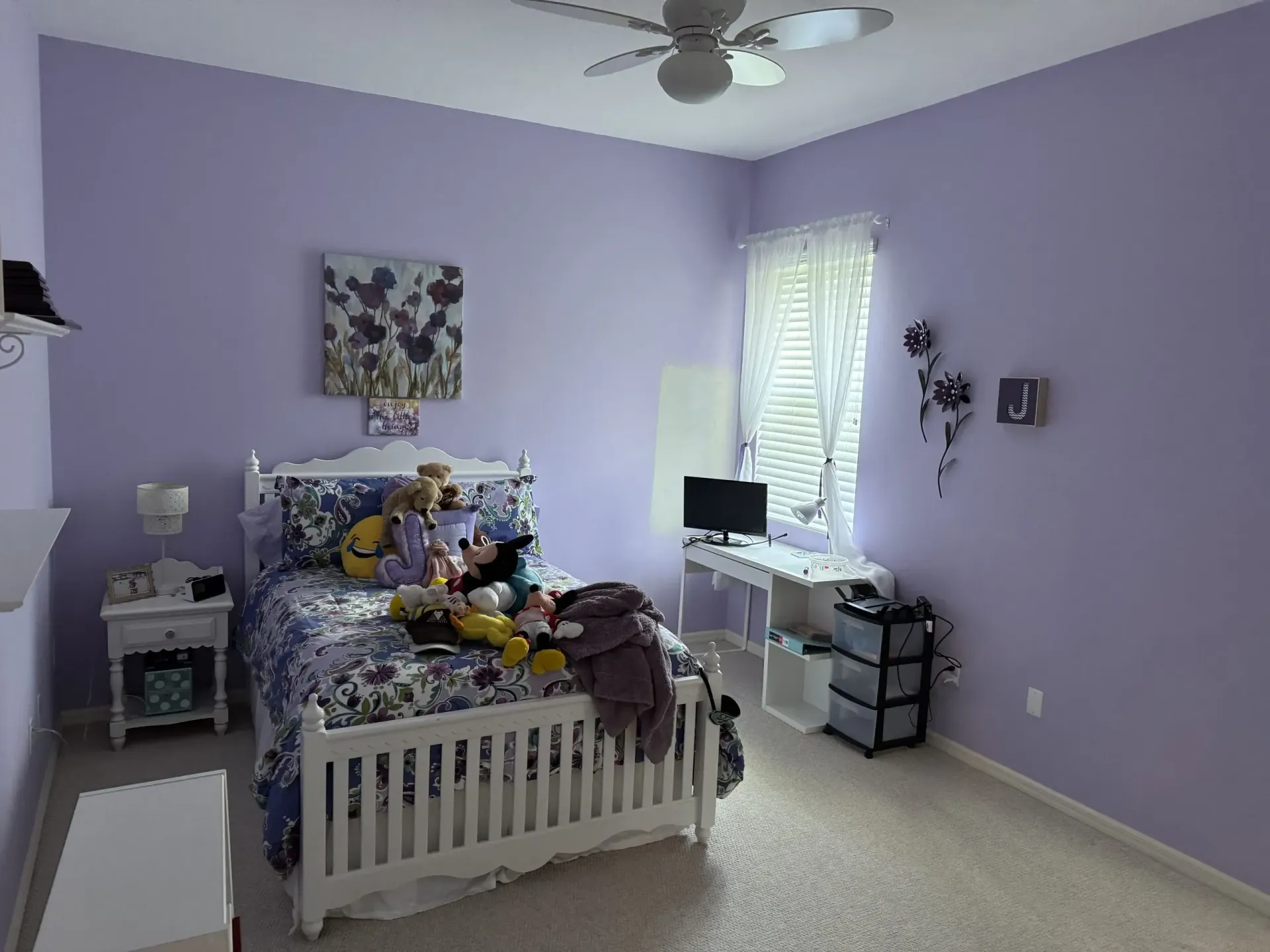Lavender bedroom with a white bed, stuffed animals, ceiling fan, and window desk area.