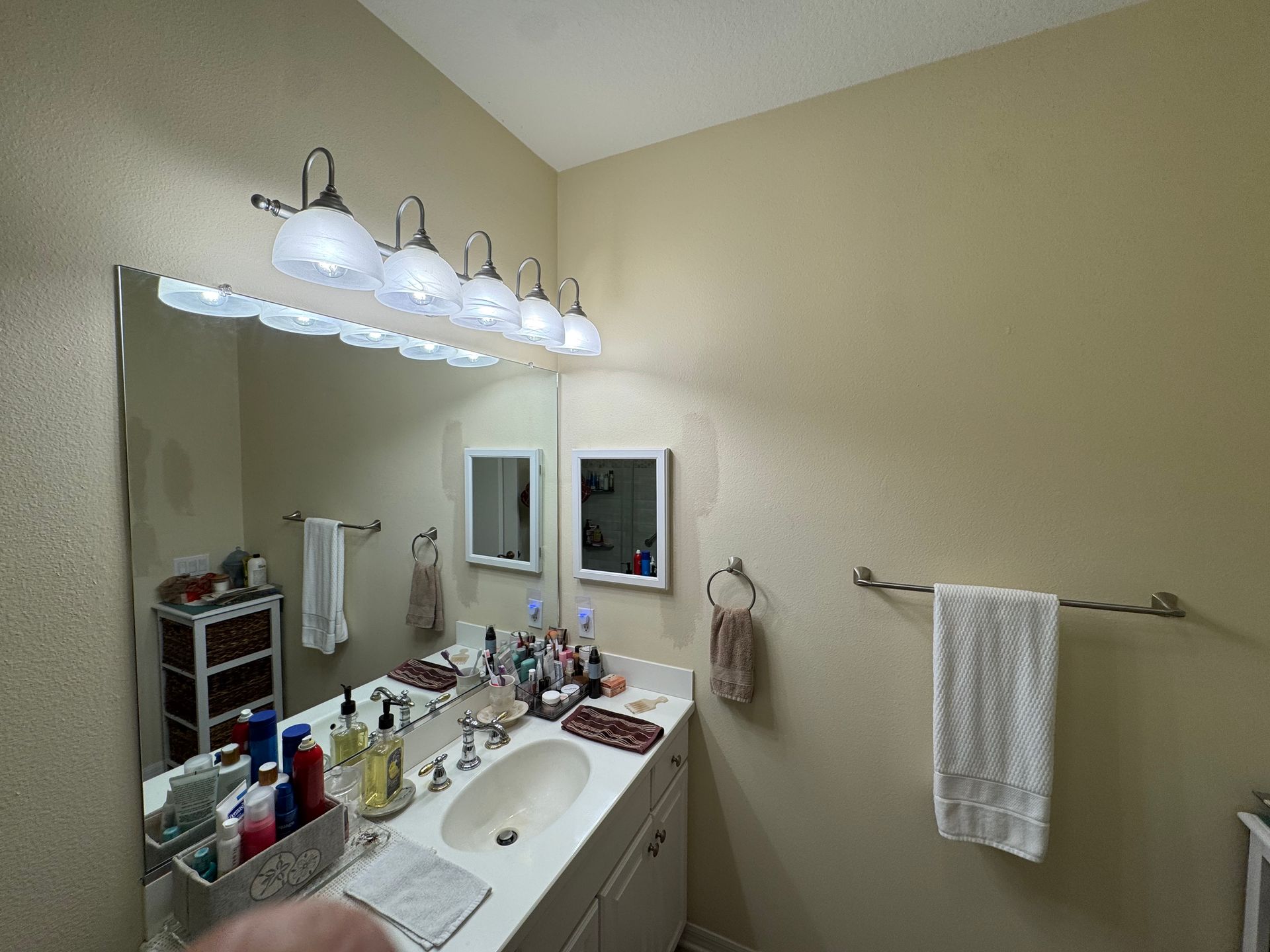 Bathroom vanity with large mirror, white sink, lights, and beige walls; toiletries clutter the counter.