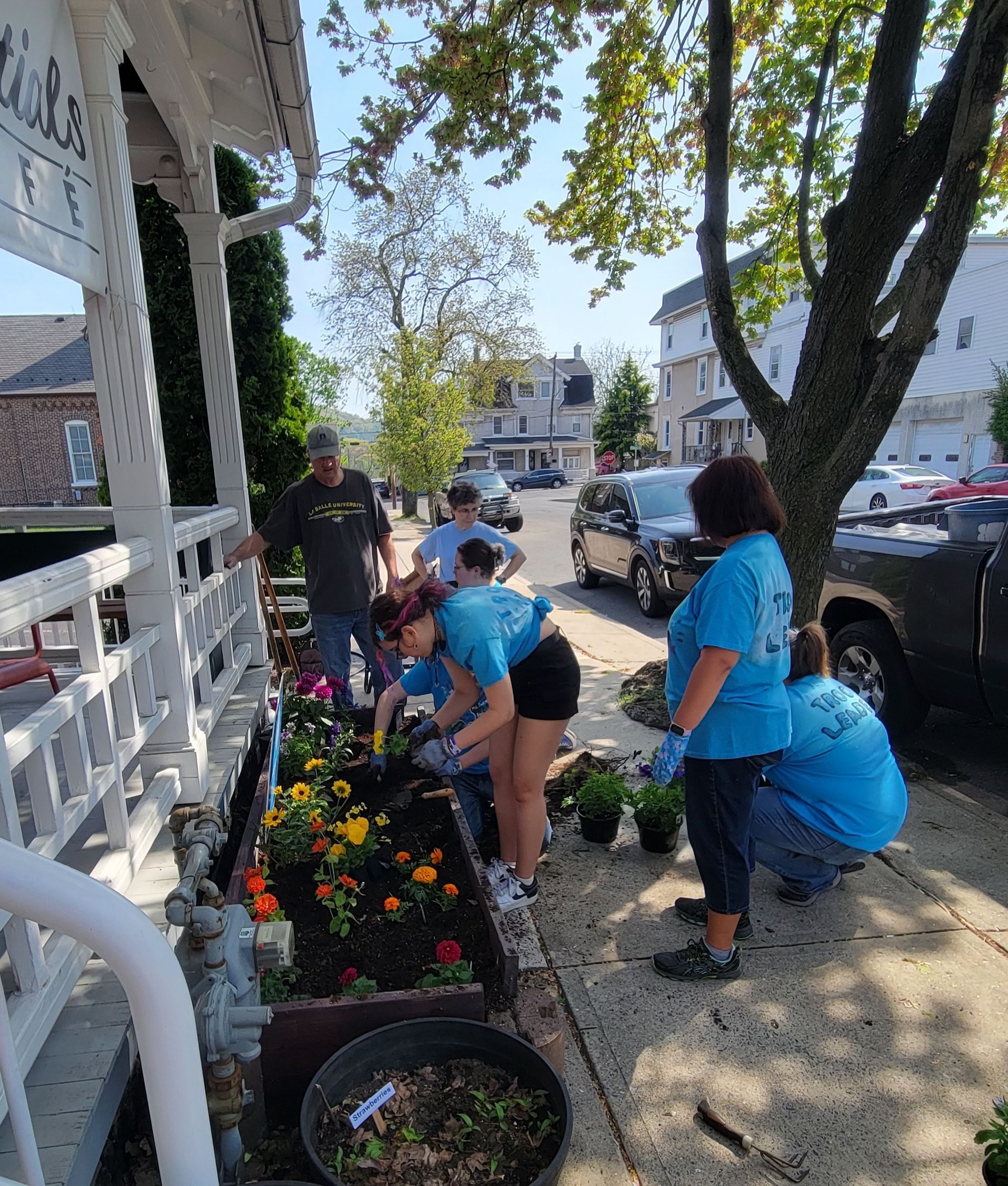 People planting flowers in a garden bed beside a white porch, on a sunny street.