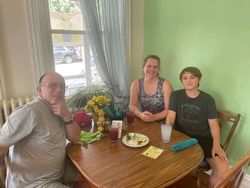 A family of three sits at a table near a window. They smile, drinks and a plate sit on the table.