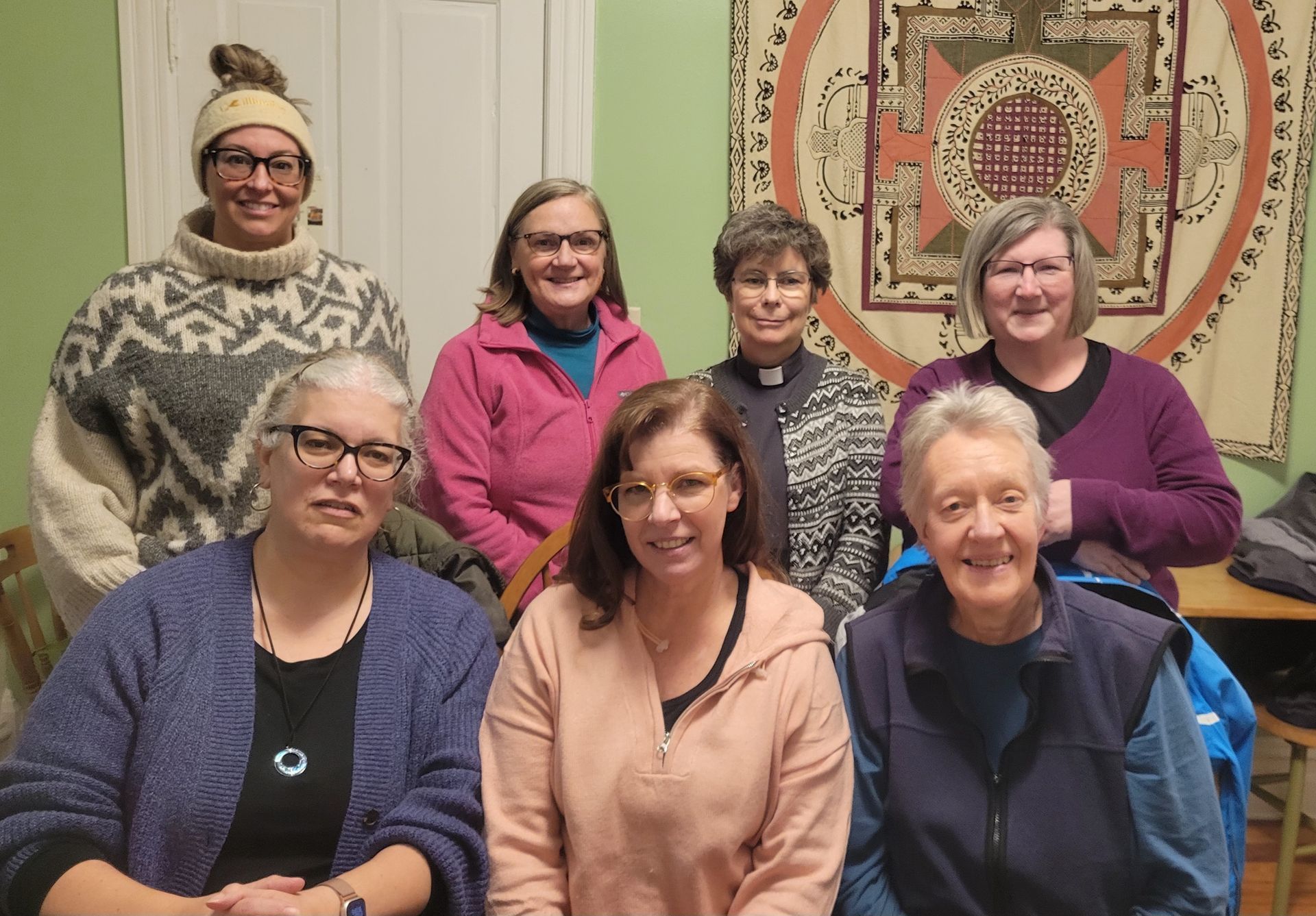 Group of seven women smiling indoors, some wearing sweaters. Tapestry background.