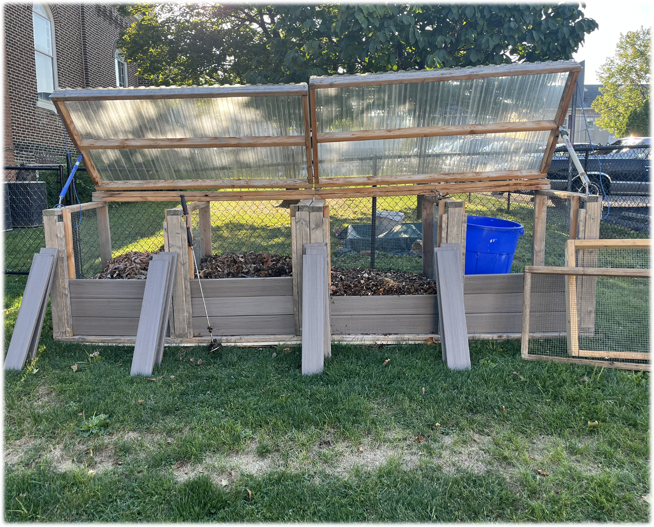 Wooden compost bins with hinged lids, on a lawn. A blue bucket sits nearby.