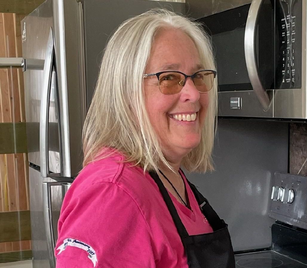 Woman with gray hair and glasses smiling in a kitchen, wearing a pink shirt and black apron.