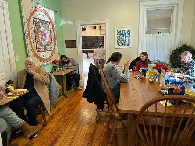People at a table and small tables in a well-lit room. Some are eating, others are talking. Decorative wall hangings.