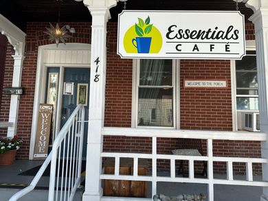 Essentials Cafe storefront with white railing, brick exterior, and a sign with a logo of leaves in a mug.