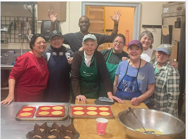 Group of people in a kitchen smile, waving near baking supplies and finished baked goods.