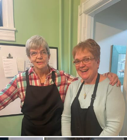 Two women, wearing aprons, smiling, and posing in a kitchen.