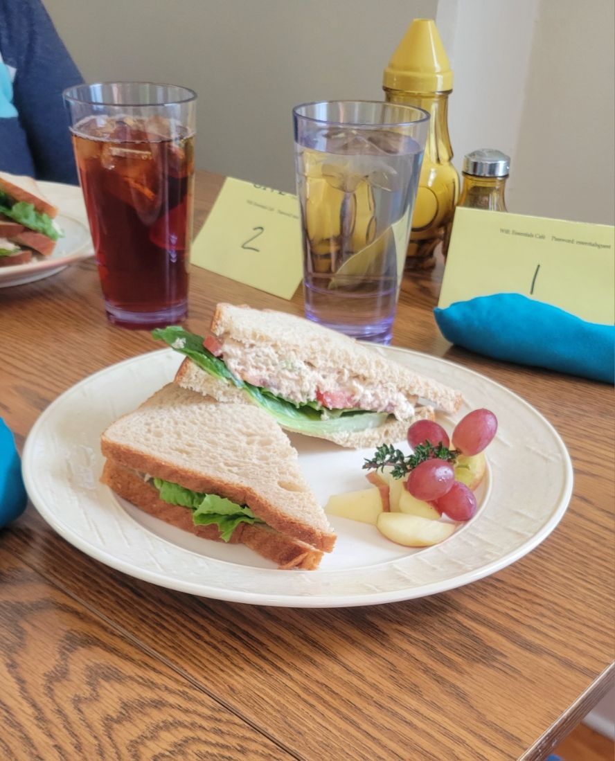 Plate with sandwich, grapes, apple slices, water, iced tea, and table setting.